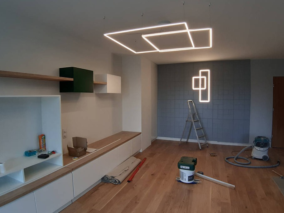 Wnętrze w trakcie prac wykończeniowych: nowoczesne oświetlenie sufitowe w kształcie kwadratów, szara ściana z geometrycznym wzorem, zabudowa meblowa z elementami drewnianymi, drabina i narzędzia...