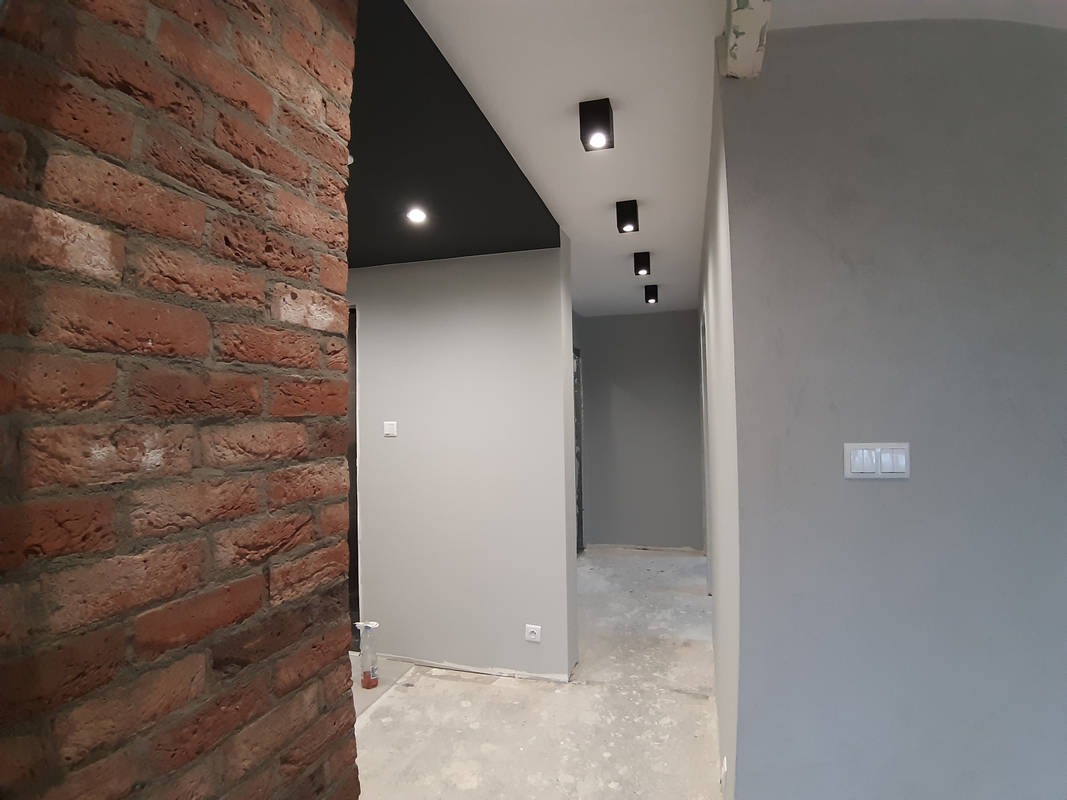 Wnętrze po remoncie: ceglana ściana, szare ściany, czarny sufit z rzędem punktowych lamp, widoczne ślady po pracach na podłodze.