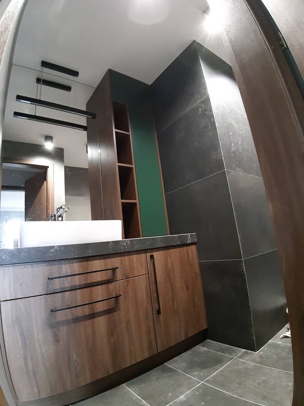 Nowoczesna łazienka z drewnianą szafką pod umywalką, czarnym blatem i wiszącą lampą liniową, uzupełniona zielonym akcentem na ścianie i ciemnymi płytkami.