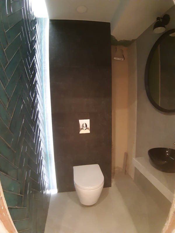 Nowoczesna łazienka w trakcie wykańczania: turkusowa ściana z płytek ułożonych w jodełkę, podświetlana krawędź, wisząca miska WC, czarna umywalka na białym blacie, okrągłe lustro z czarną ramą.