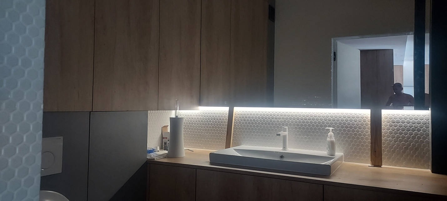 Nowoczesna łazienka z umywalką nablatową, podświetlanym lustrem i heksagonalnymi płytkami na ścianie; w lustrze odbija się sylwetka osoby.