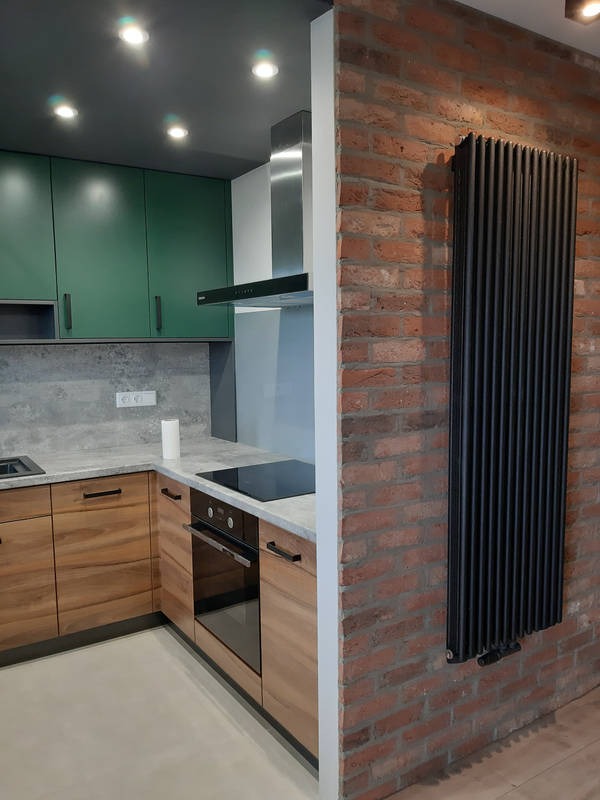 Nowoczesna kuchnia z drewnianymi szafkami, zielonymi górnymi szafkami, szarym blatem i ceglaną ścianą z czarnym grzejnikiem żeberkowym.