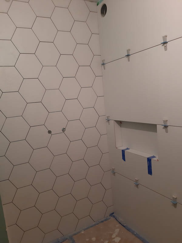 Ściana łazienki w trakcie układania białych, heksagonalnych płytek. Widoczne krzyżyki dystansowe i poziomica sznurkowa. Przygotowana wnęka na półkę.