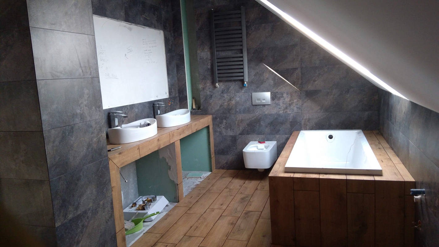 Łazienka na poddaszu w trakcie wykańczania: drewnopodobne płytki na podłodze, obudowa wanny i umywalek z surowego drewna, szare płytki na ścianach, biała wanna i umywalki, widoczny fragment...