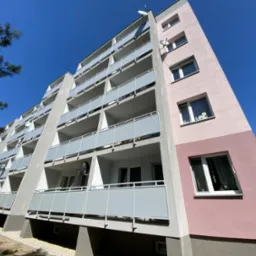 Wielopiętrowy budynek mieszkalny po renowacji z balkonami z szarymi barierkami, świeżo pomalowaną elewacją w odcieniach szarości i różu, widoczny z dołu pod ostrym kątem w słoneczny dzień.