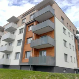 Nowoczesny budynek wielorodzinny z elewacją w kolorach: białym, beżowym i imitującym drewno, z balkonami o ażurowych balustradach i widocznym trawnikiem.