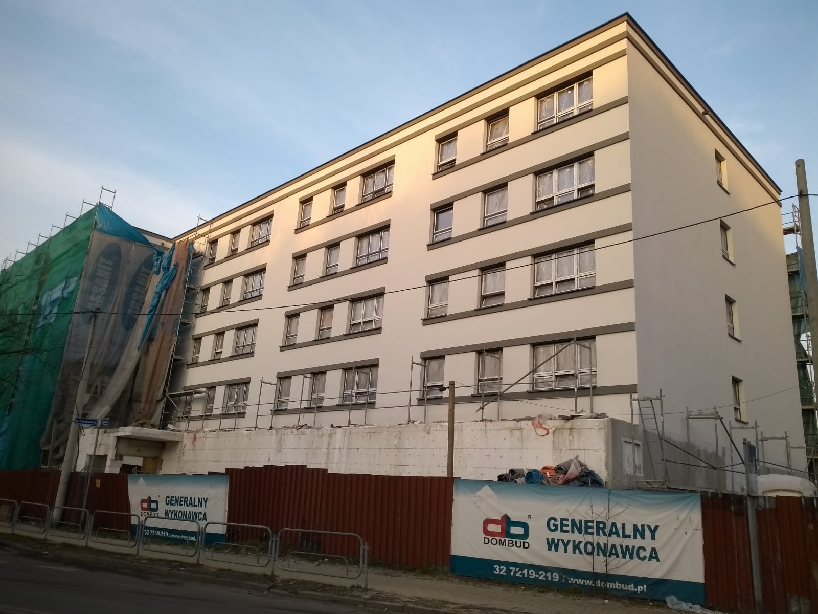Wielopiętrowy budynek po renowacji elewacji, widoczne rusztowania z boku budynku i banery reklamowe wykonawcy u dołu, częściowo zakryte ogrodzeniem.
