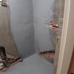 Osoba prywatna - Przygotowanie ścian i podłogi w łazience do dalszych prac wykończeniowych; widoczne ślady po instalacjach i zabezpieczona szara powłoka hydroizolacyjna.