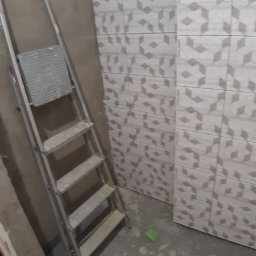 Osoba prywatna - Ściana w trakcie układania płytek w łazience, widoczna drabina malarska i fragment podłogi z pozostałościami zaprawy.