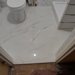 Osoba prywatna - Biała toaleta i brodzik z białymi płytkami imitującymi marmur, widoczne rury instalacyjne i fragment podłogi z brązowym materiałem zabezpieczającym.