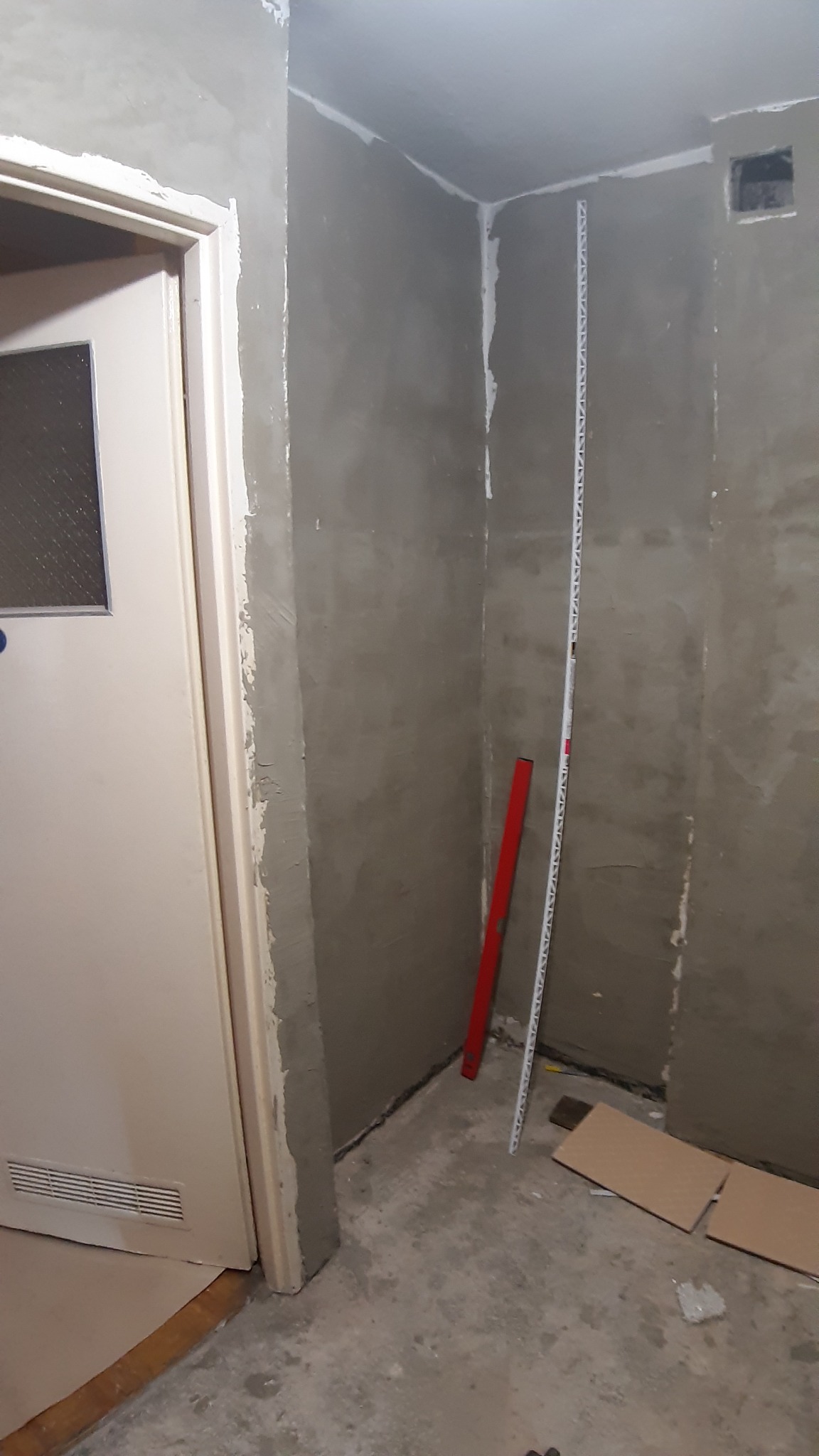 Ściany w trakcie szpachlowania z widocznym pionem i poziomnicą, zabezpieczona podłoga, surowy stan wykończenia.