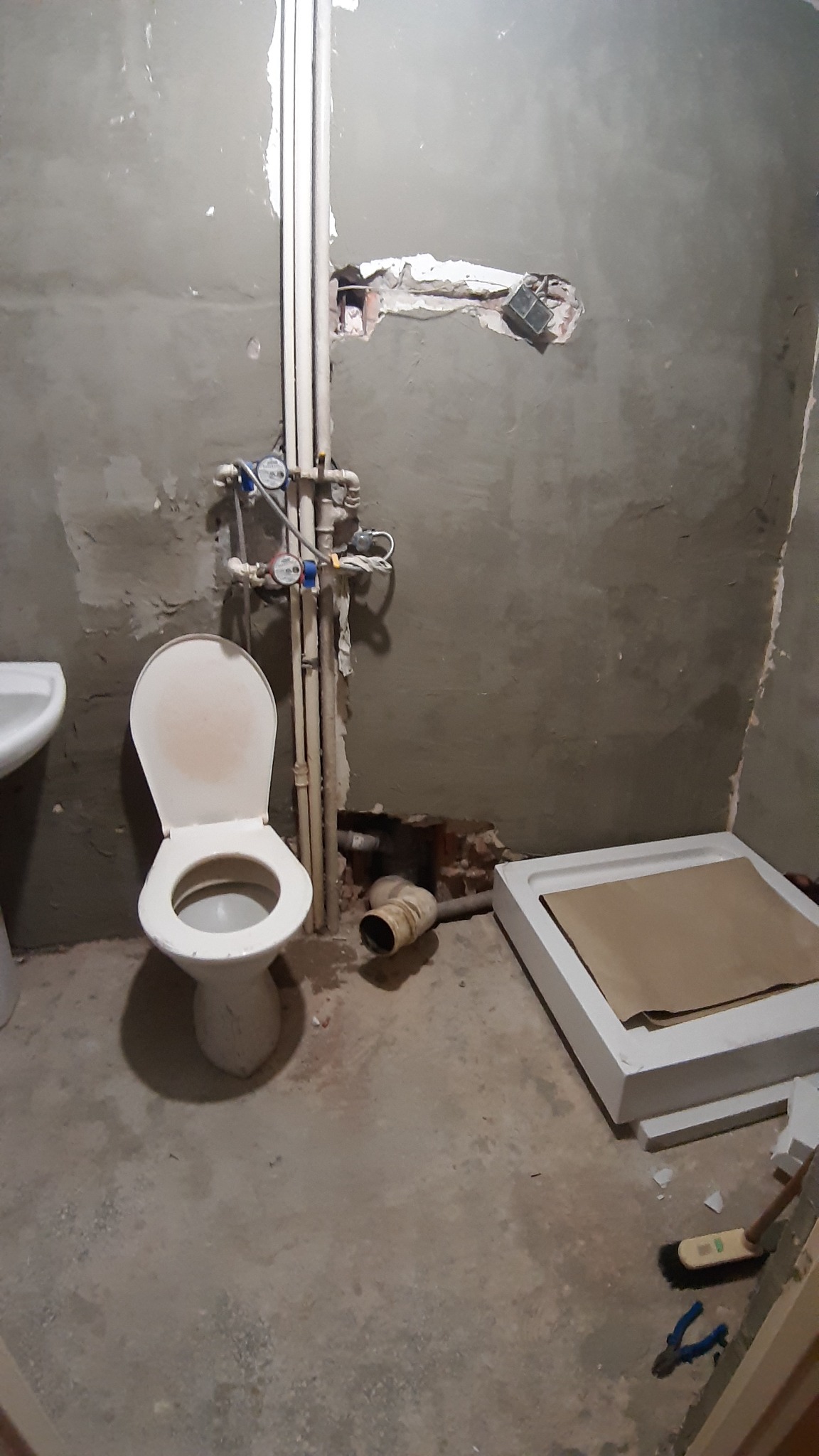 Łazienka w trakcie remontu: surowe ściany, niezabudowane rury, toaleta z otwartą klapą, brodzik zabezpieczony kartonem, widoczne narzędzia i gruz.