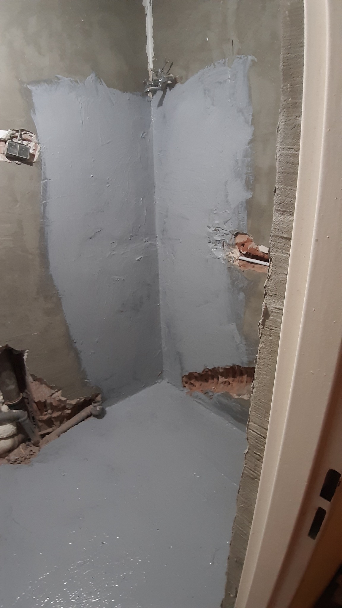 Przygotowanie ścian i podłogi w łazience do dalszych prac wykończeniowych; widoczne ślady po instalacjach i zabezpieczona szara powłoka hydroizolacyjna.