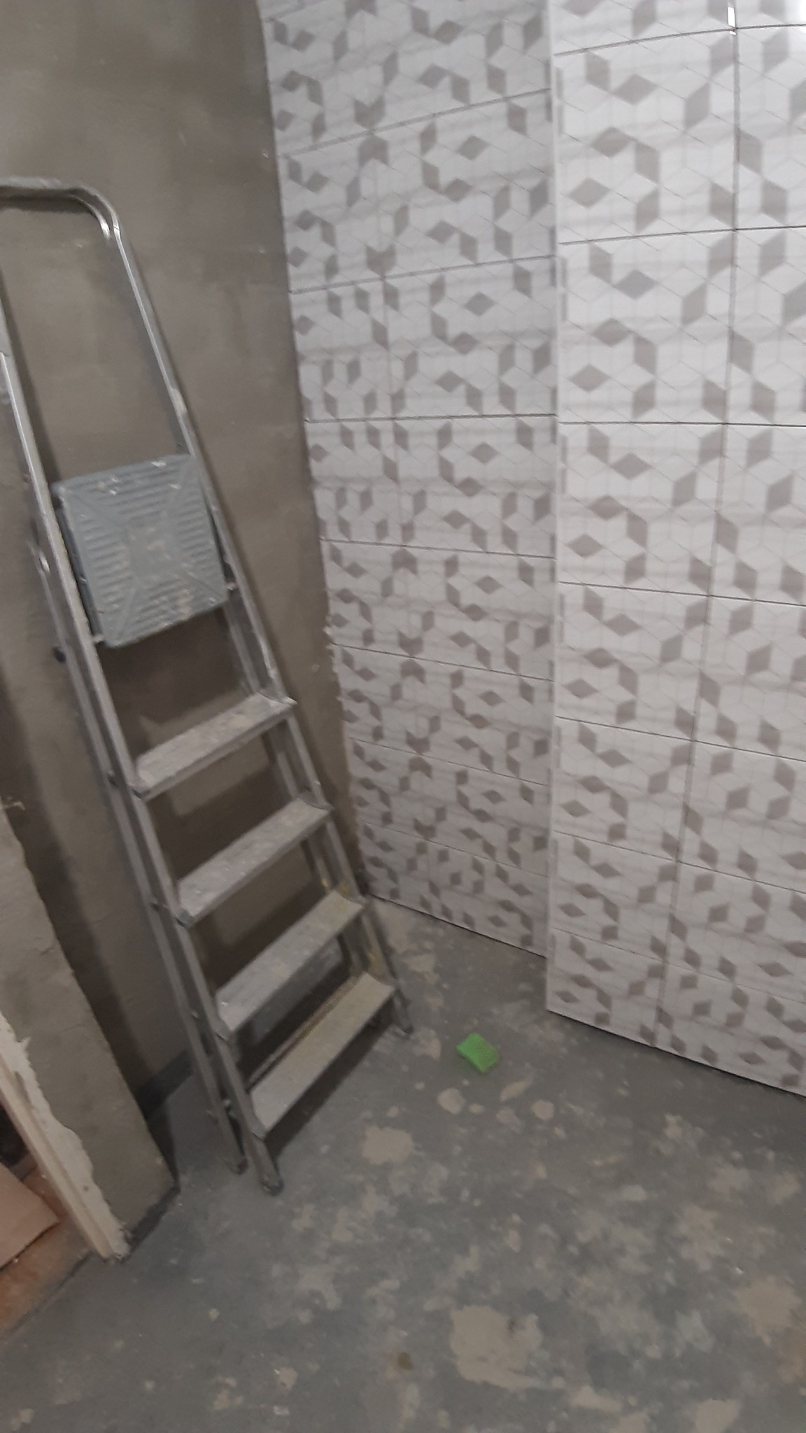 Ściana w trakcie układania płytek w łazience, widoczna drabina malarska i fragment podłogi z pozostałościami zaprawy.