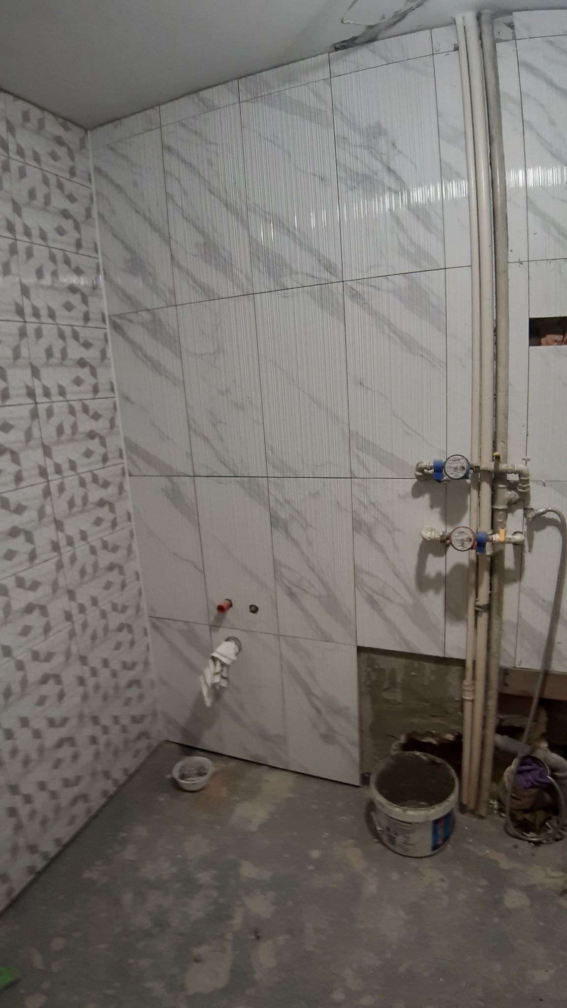 Łazienka w trakcie remontu: częściowo wyłożone płytki imitujące marmur na ścianach, odsłonięte rury z licznikami, widoczny fragment nieukończonej ściany i wiadro z zaprawą na podłodze.