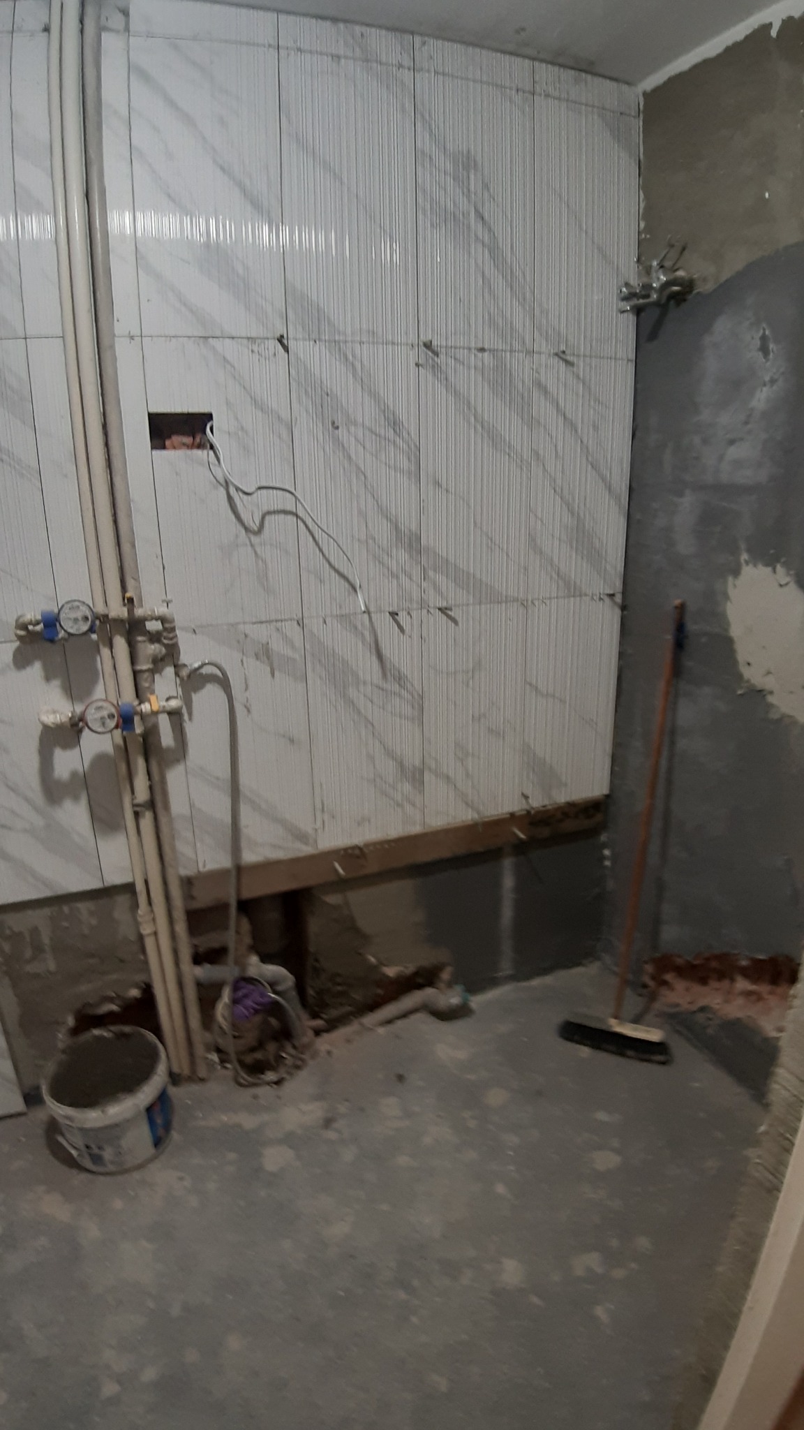 Łazienka w trakcie prac: częściowo ułożone białe płytki z delikatnym wzorem na ścianie, widoczne rury, instalacja wodna, fragment surowej ściany, szczotka do zamiatania i wiadro z resztkami zaprawy...