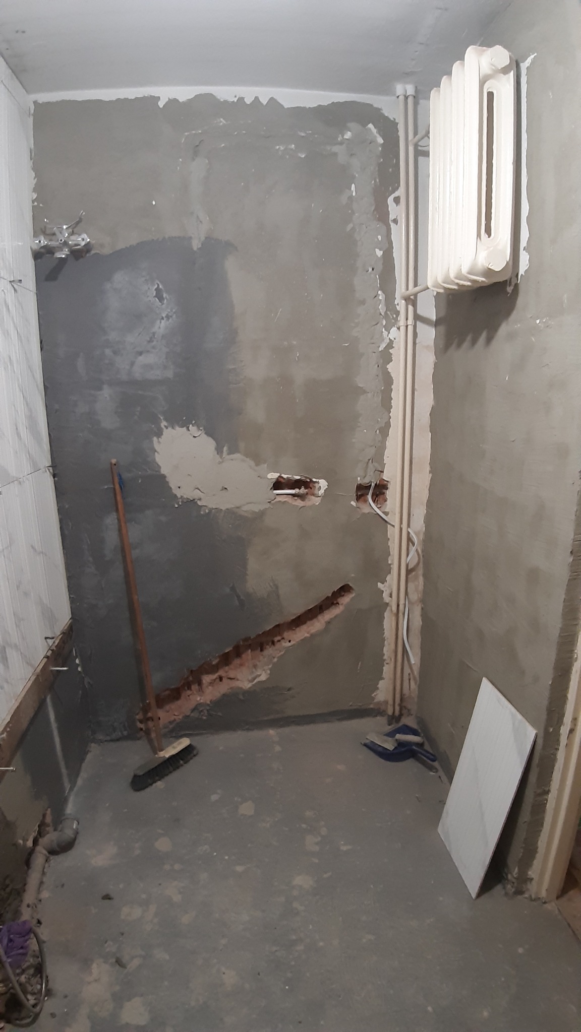 Łazienka w trakcie prac remontowych: surowe ściany z widocznymi śladami po skuciu, grzejnik na ścianie, rury, szczotka i płytka ceramiczna na podłodze, widoczne instalacje elektryczne.