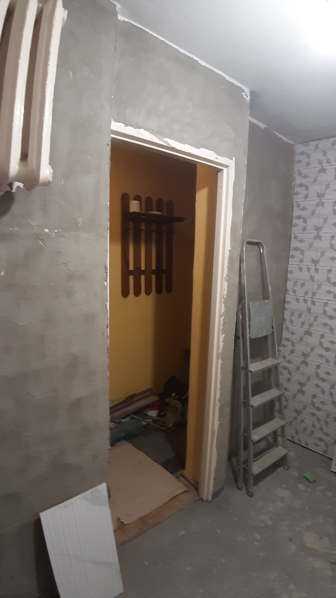 Ściana w trakcie gruntowania pod malowanie, widoczna drabina, grzejnik i fragment wykończonej łazienki z płytkami wzorzystymi.