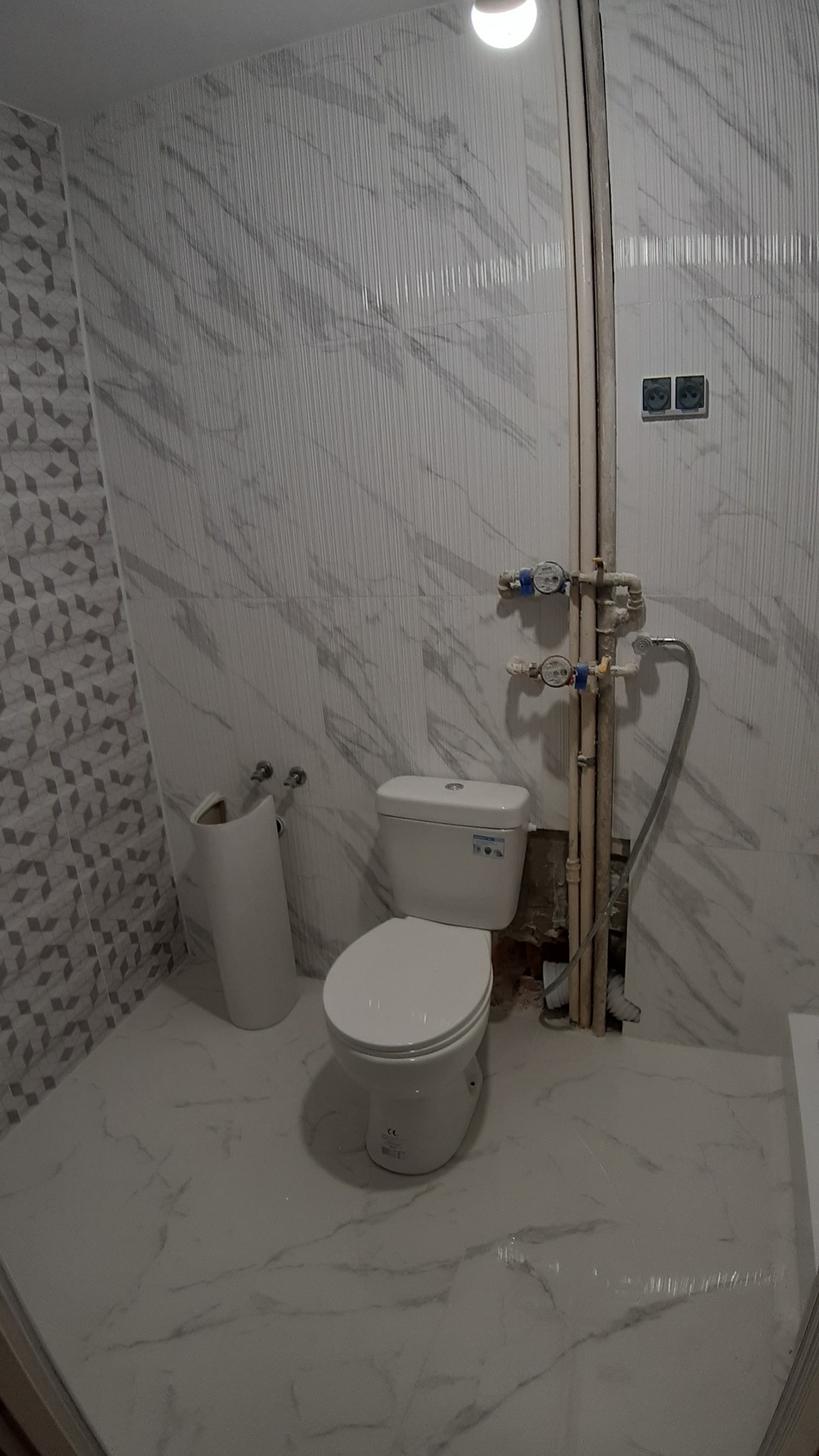 Biała toaleta kompaktowa z zamontowanymi licznikami wody na tle marmurowych płytek ściennych w trakcie remontu łazienki.