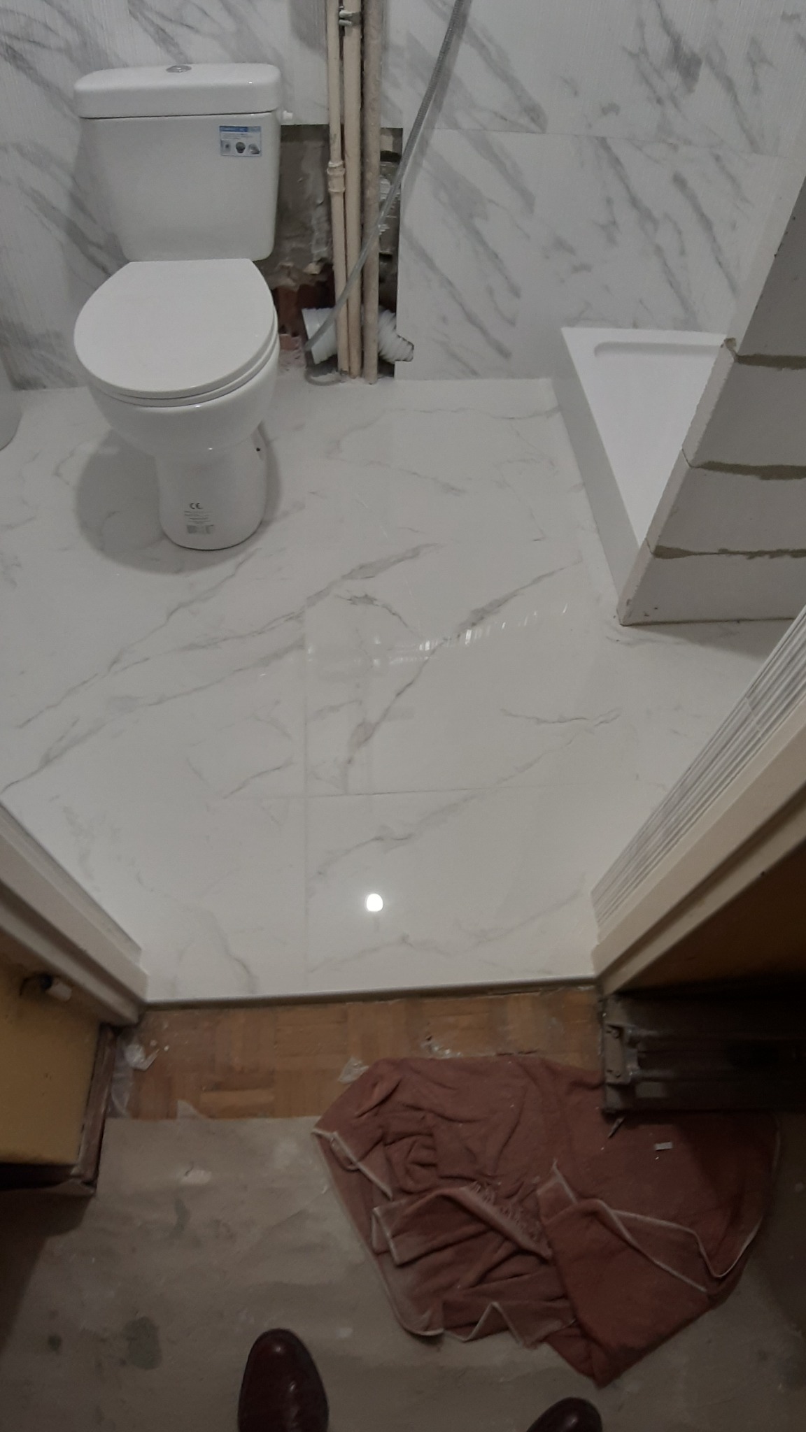 Biała toaleta i brodzik z białymi płytkami imitującymi marmur, widoczne rury instalacyjne i fragment podłogi z brązowym materiałem zabezpieczającym.