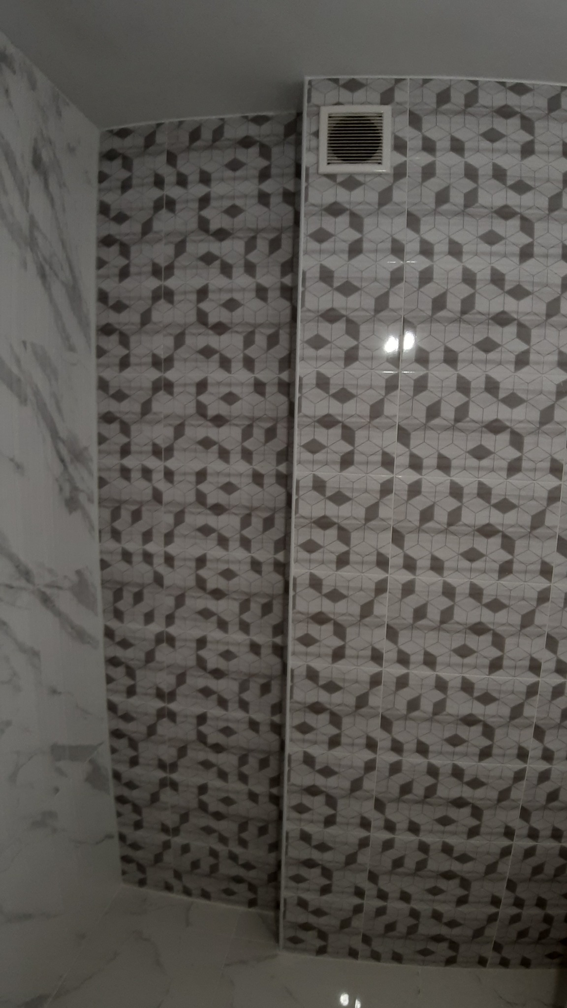 Fragment wykończonej łazienki z szaro-białymi płytkami w geometryczne wzory na ścianach i podłodze, widoczna kratka wentylacyjna.