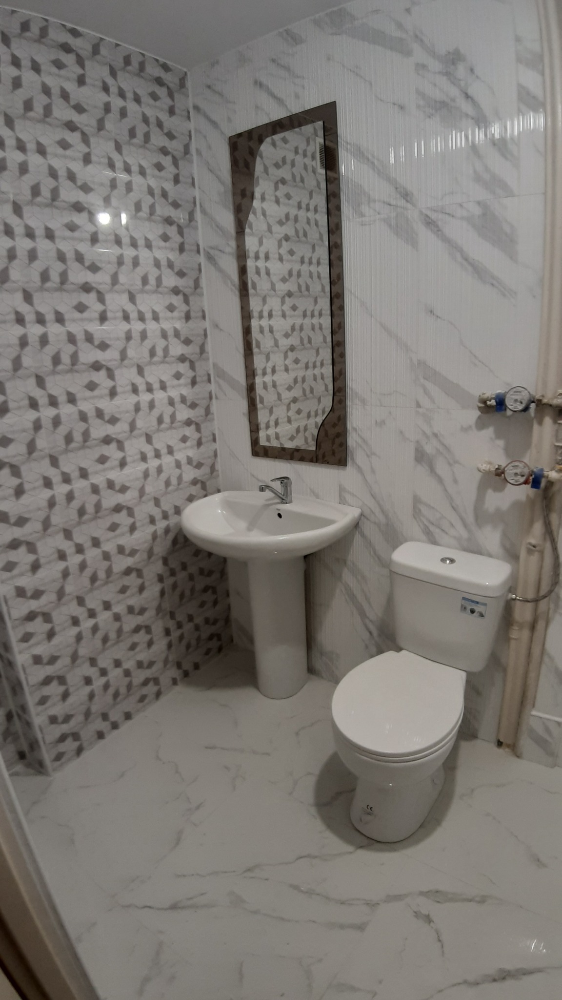 Umywalka na postumencie, toaleta i lustro w ramie na tle ściany z płytek o wzorze geometrycznym i marmurowych, w wykończonej łazience.