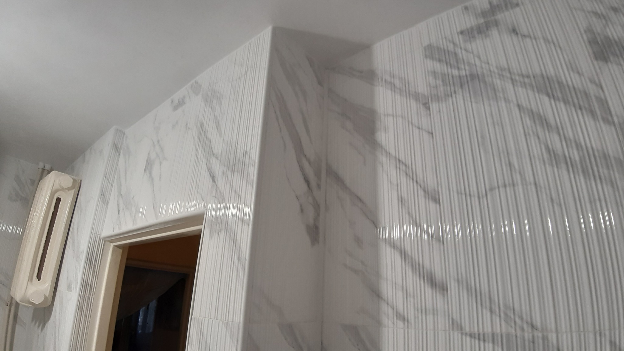 Wykończona łazienka z białymi płytkami imitującymi marmur z delikatnymi pionowymi żłobieniami, widoczny fragment grzejnika i ościeżnica drzwi.