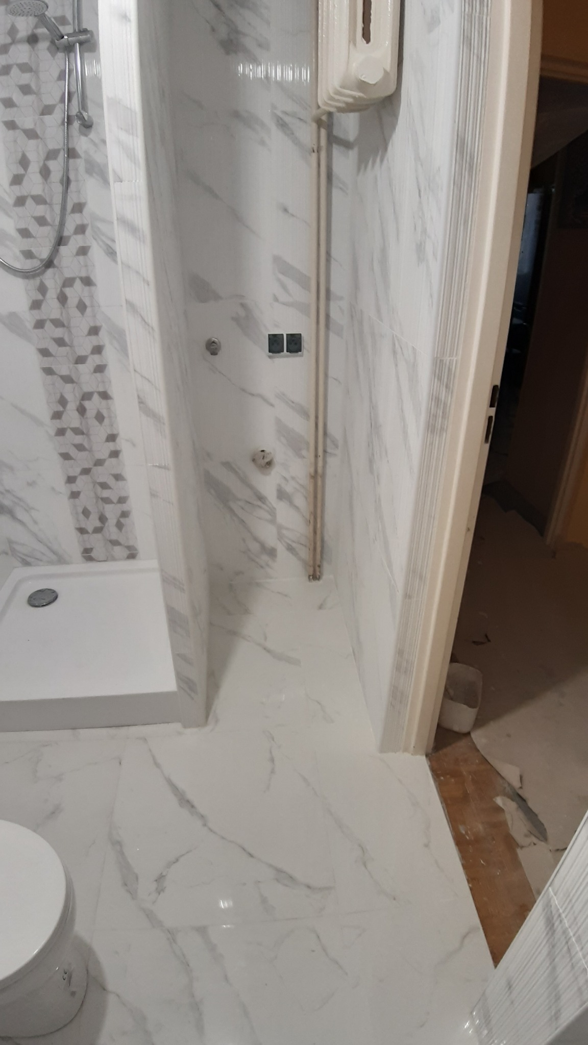 Wykończona kabina prysznicowa z białymi płytkami imitującymi marmur, widoczny brodzik, bateria, rury, dozownik mydła oraz fragment toalety i drzwi.