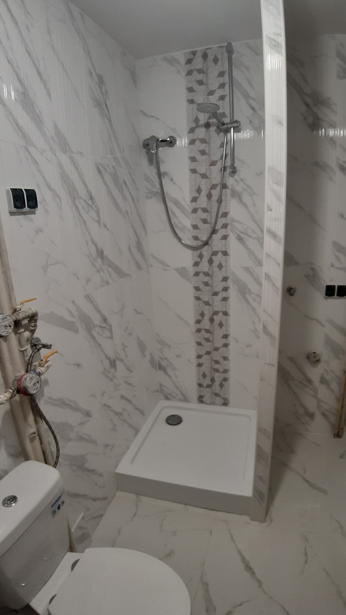 Nowoczesna łazienka z biało-szarymi marmurkowymi płytkami, kabiną prysznicową z geometrycznym akcentem, toaletą i instalacją wodną z licznikami.