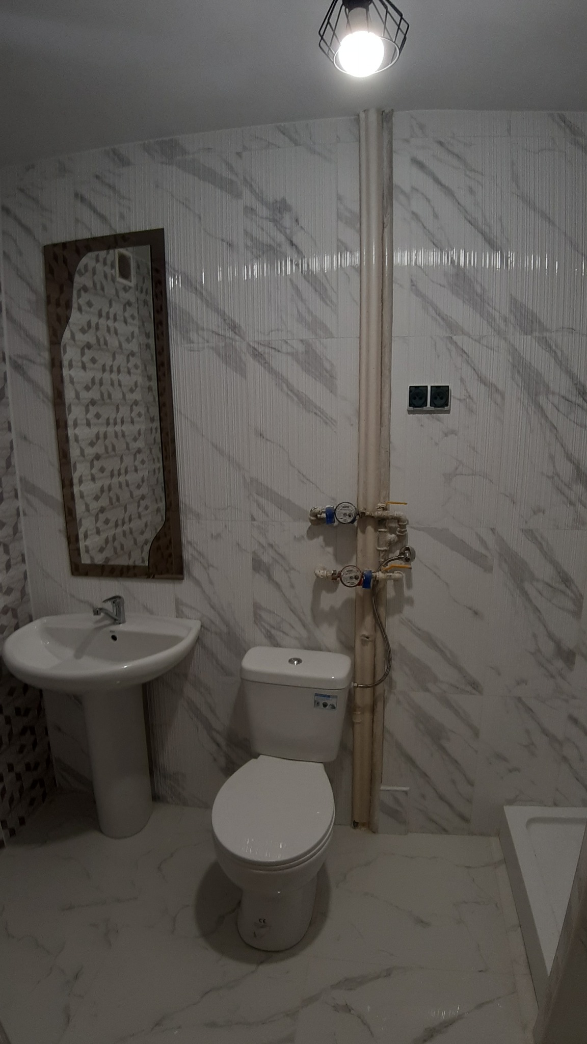 Nowoczesna łazienka z marmurowymi płytkami, wiszącą umywalką, toaletą i czarną lampą sufitową w geometrycznej obudowie.