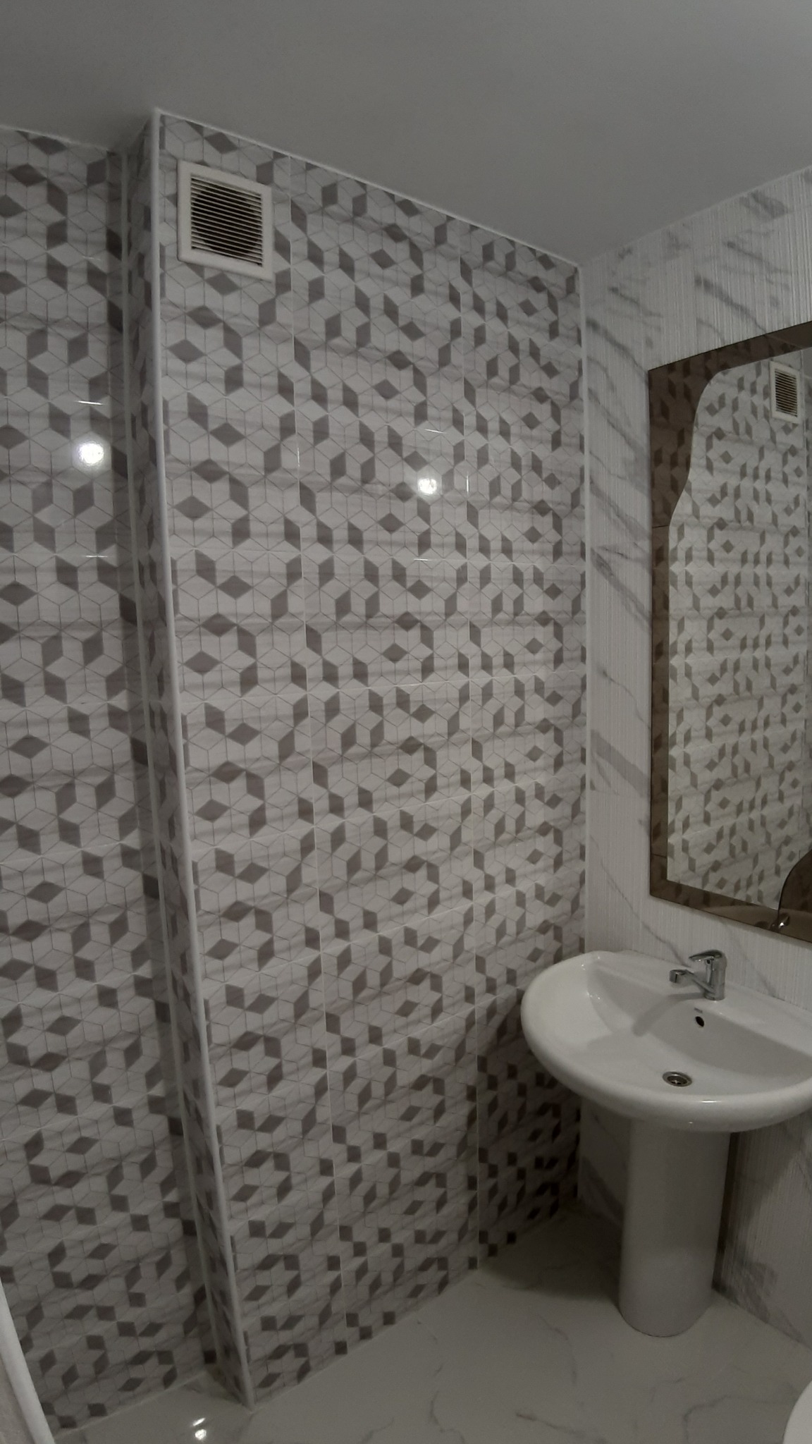 Nowoczesna łazienka z umywalką na postumencie, ściany wyłożone szaro-białymi płytkami z geometrycznym wzorem kostek, widoczny fragment lustra i kratka wentylacyjna.