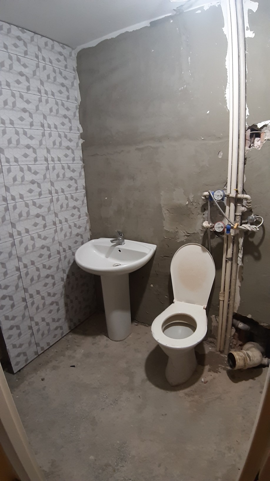 Łazienka w trakcie prac wykończeniowych: częściowo ułożone płytki na ścianie, surowy tynk, zamontowana umywalka na postumencie i toaleta przed podłączeniem, widoczne rury i liczniki.