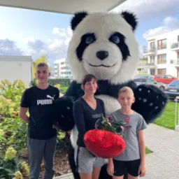 Uśmiechnięta rodzina pozuje z animatorem w stroju pandy trzymającym balon w kształcie serca na tle nowoczesnego osiedla