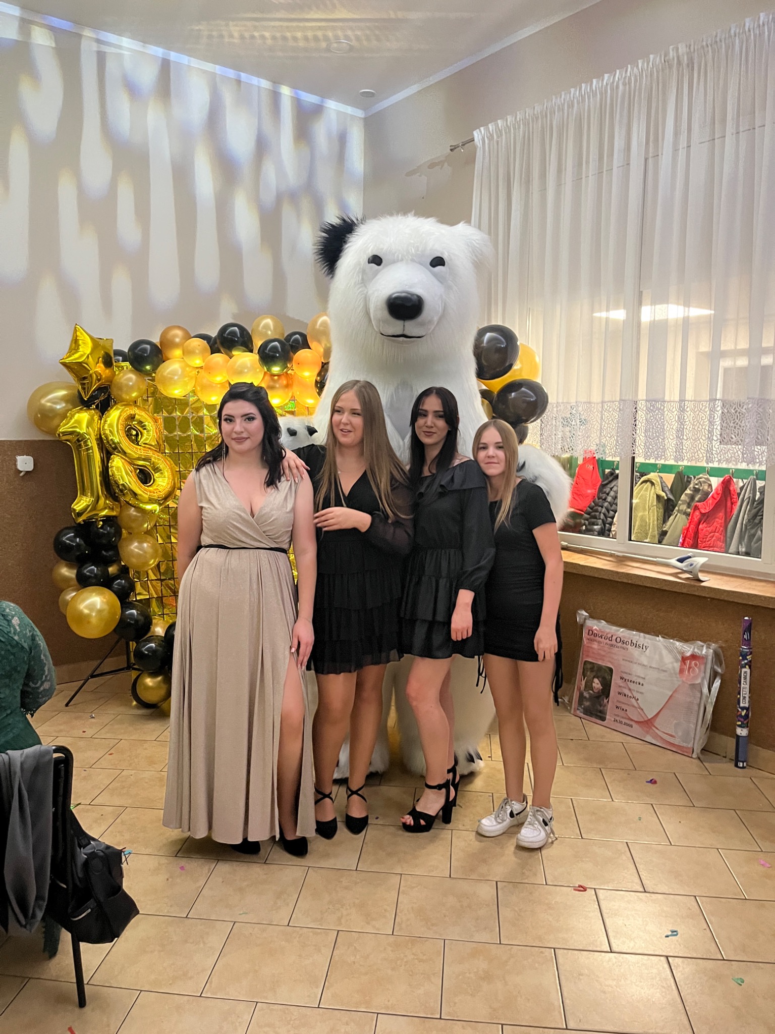 Cztery młode kobiety pozują do zdjęcia z dużym pluszowym białym niedźwiedziem na tle dekoracji z czarno-złotych balonów i cyfrą '18' z balonów.