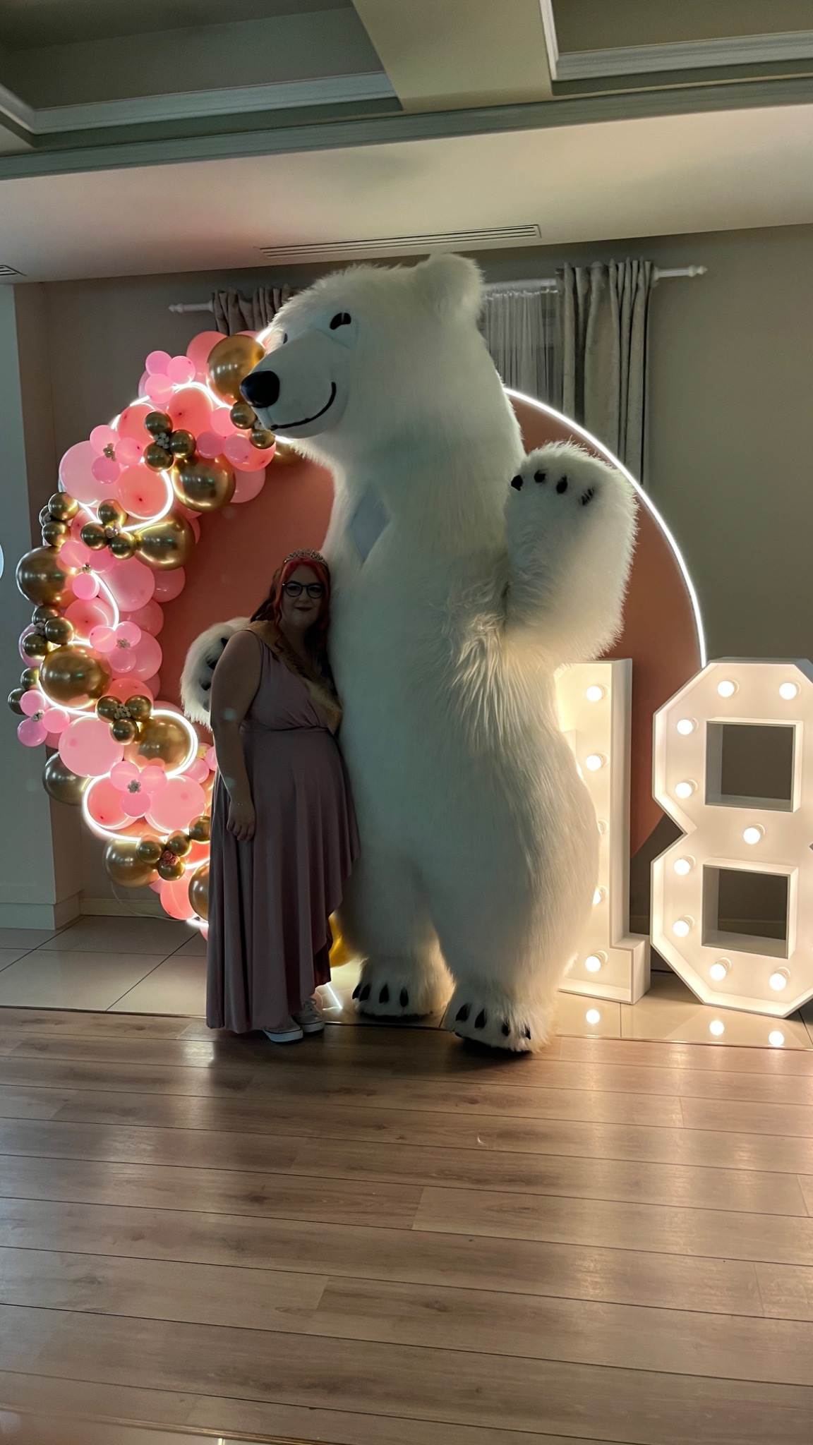 Kobieta w sukience pozuje obok dużego, białego misia w kostiumie, na tle dekoracji z różowych i złotych balonów oraz świecących cyfr '18'.