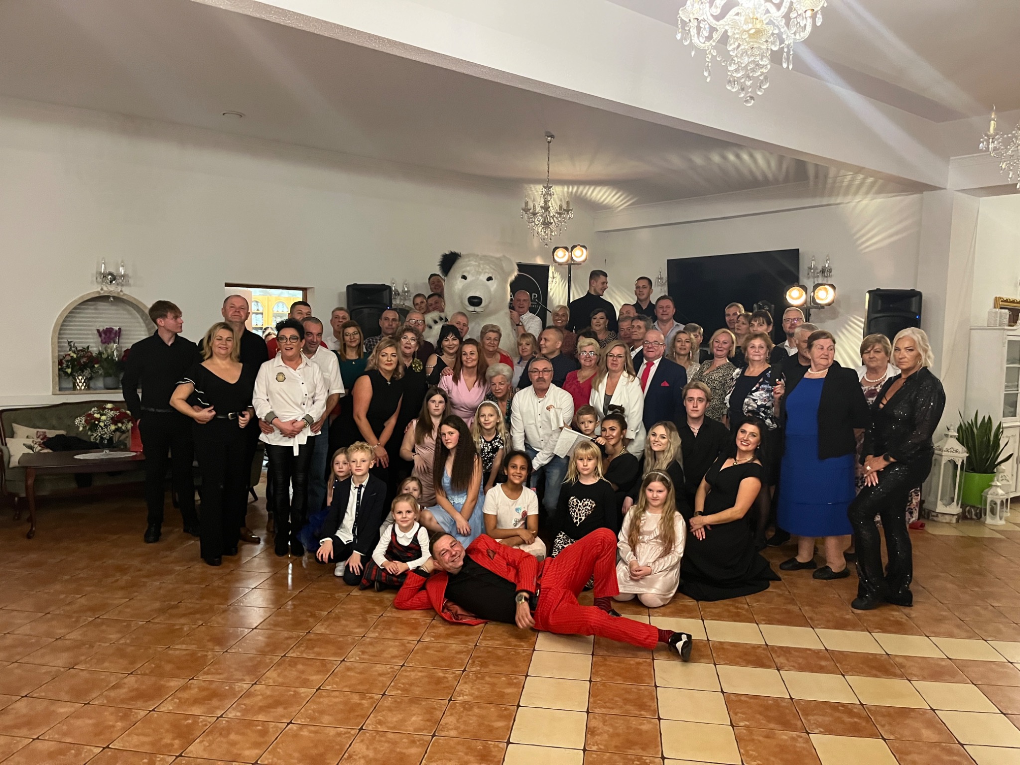 Grupowe zdjęcie uczestników eventu w Gdańsku, z mężczyzną w czerwonym garniturze leżącym na podłodze i osobą w kostiumie białego misia w tle.