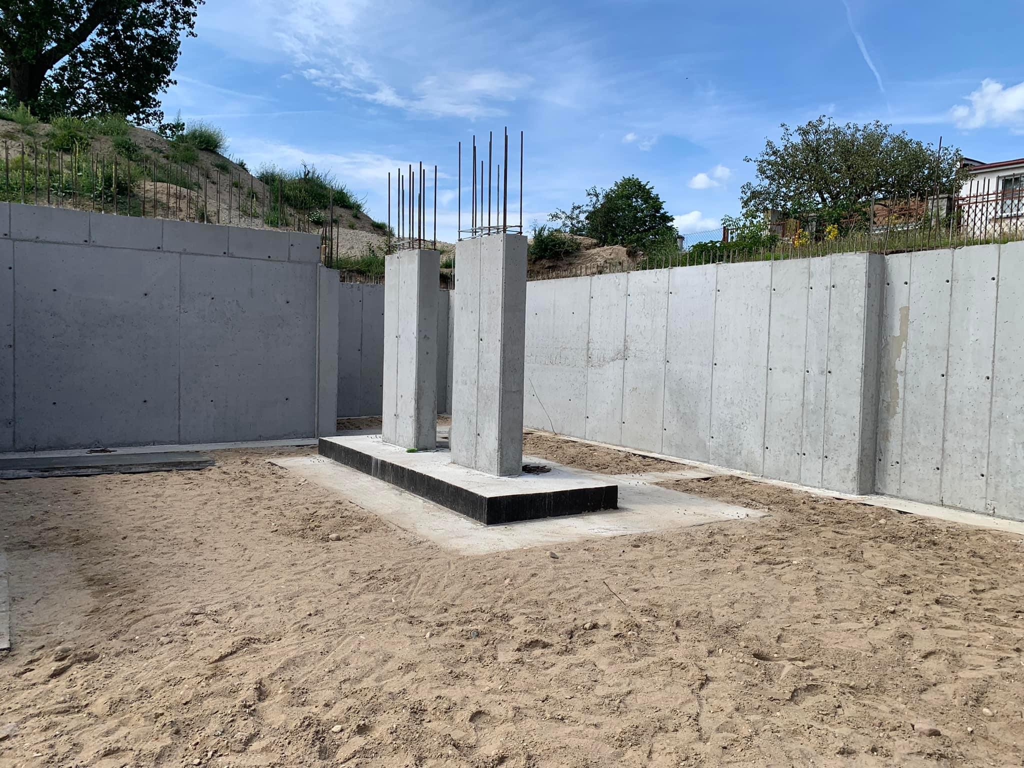 Betonowe fundamenty z wystającymi prętami zbrojeniowymi, otoczone ścianami oporowymi, na piaszczystym terenie budowy w słoneczny dzień.