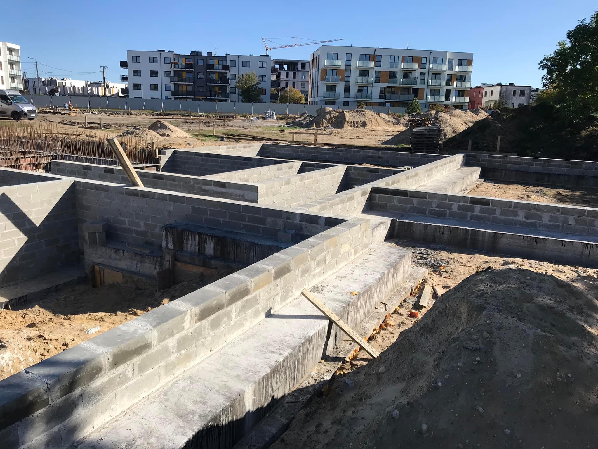 Wykop pod fundamenty z częściowo wzniesionymi ścianami z bloczków betonowych, na tle zabudowy wielorodzinnej i błękitnego nieba.