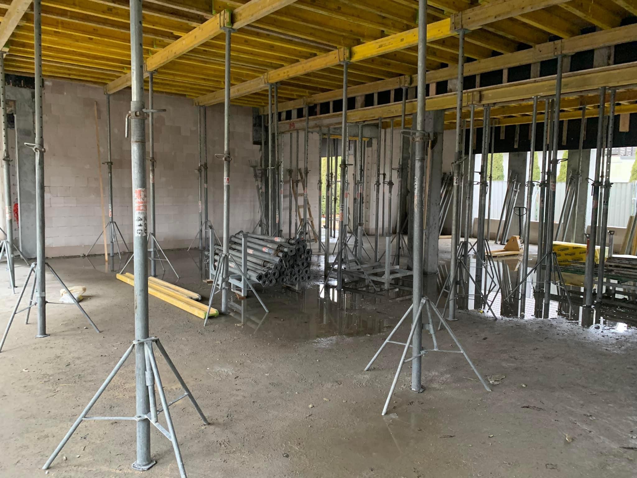 Wnętrze murowanego domu w trakcie budowy z widocznymi podporami stropowymi, mokrą posadzką i częściowo wymurowanymi ścianami z bloczków.