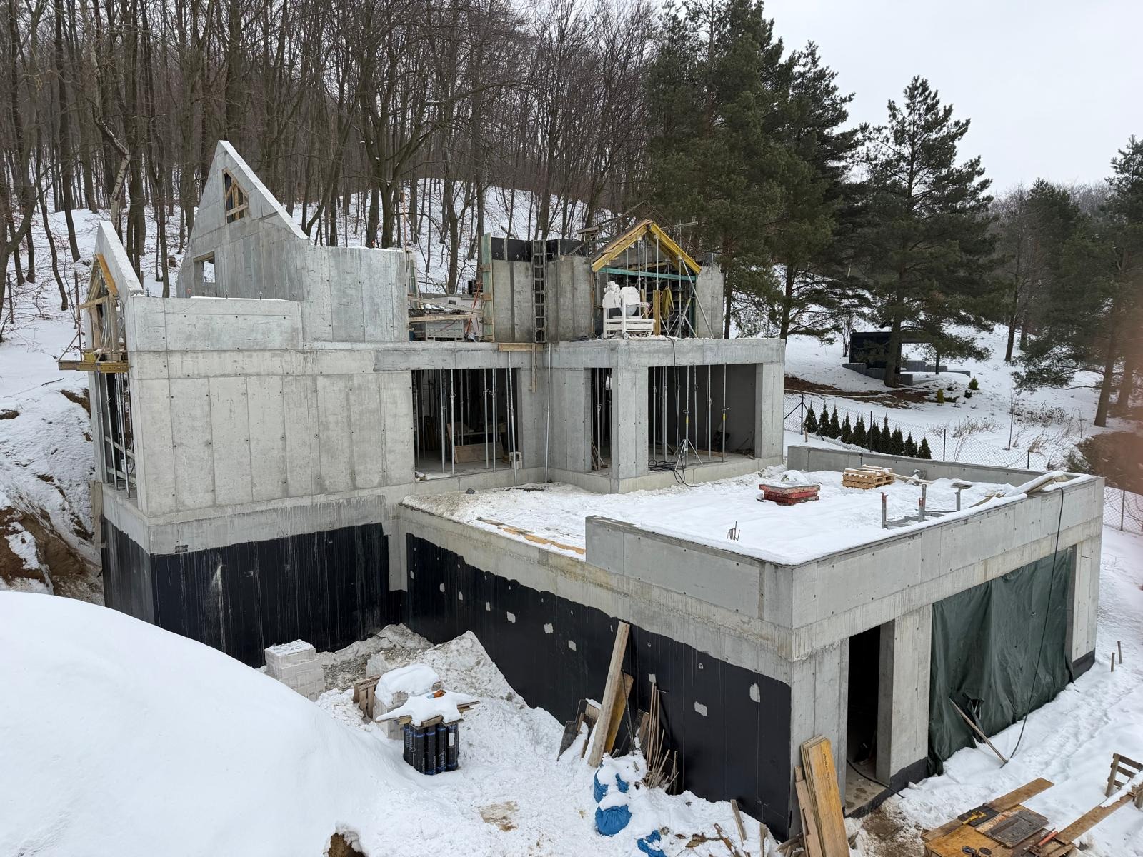 Surowy stan domu z betonu w zimowej scenerii. Widoczne szalunki, zbrojenia i izolacja fundamentów. Częściowo zadaszona konstrukcja w Redzie.