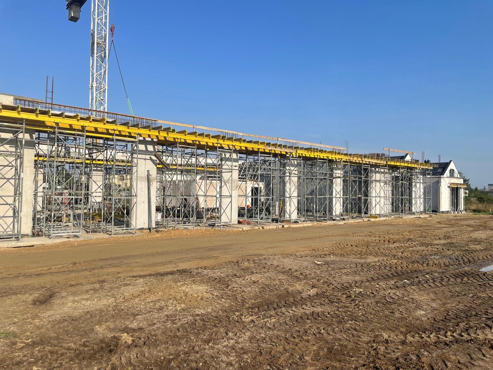 Wznoszenie konstrukcji budynku z betonu i stali, widoczne szalunki i zbrojenia. W tle fragment gotowego budynku i żuraw budowlany na tle błękitnego nieba.