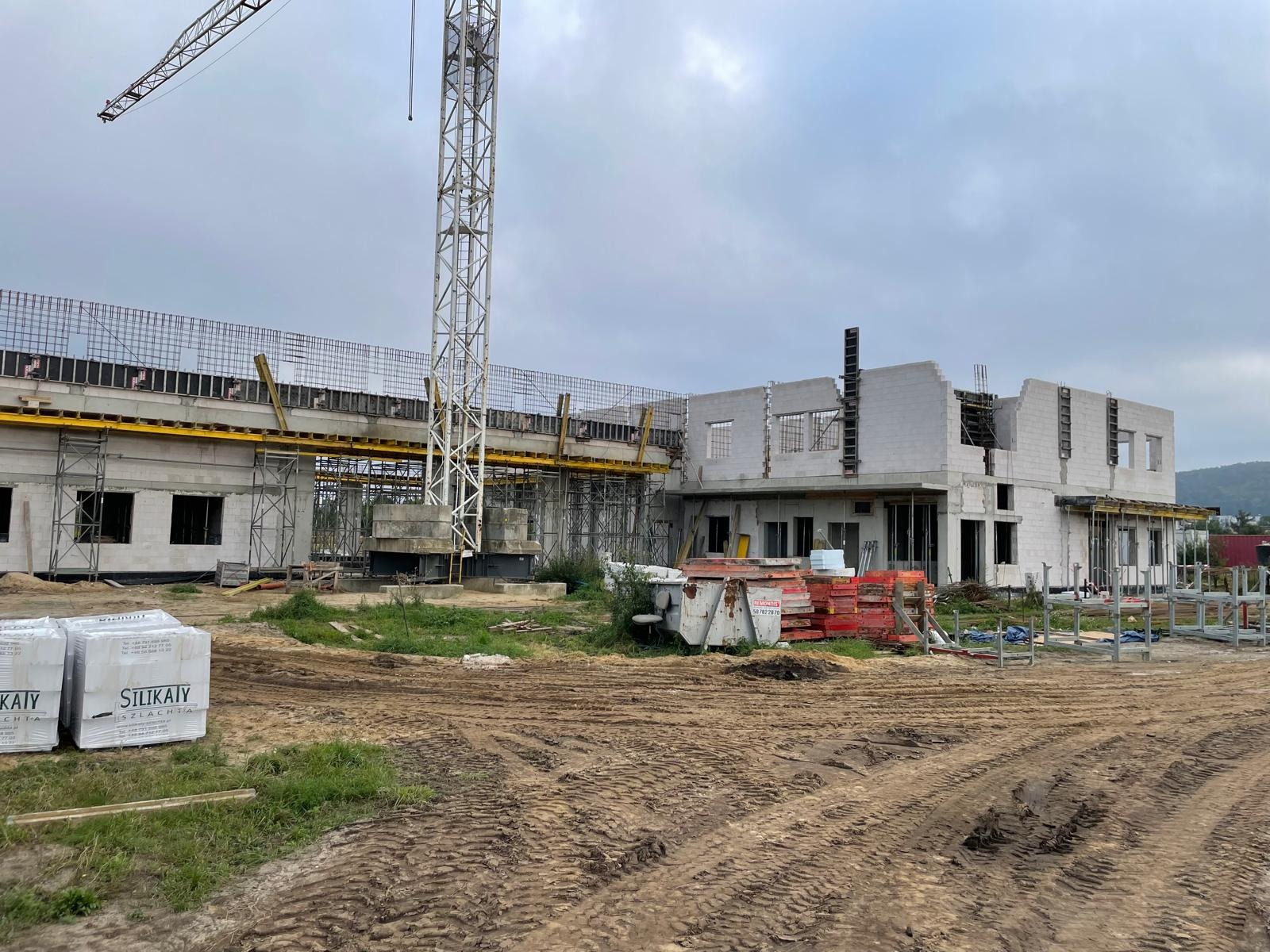 Budowa hali w toku: widoczne ściany z betonu komórkowego, zbrojenia, szalunki, dźwig. Na pierwszym planie palety z bloczkami SILKA. Teren budowy, pochmurne niebo.