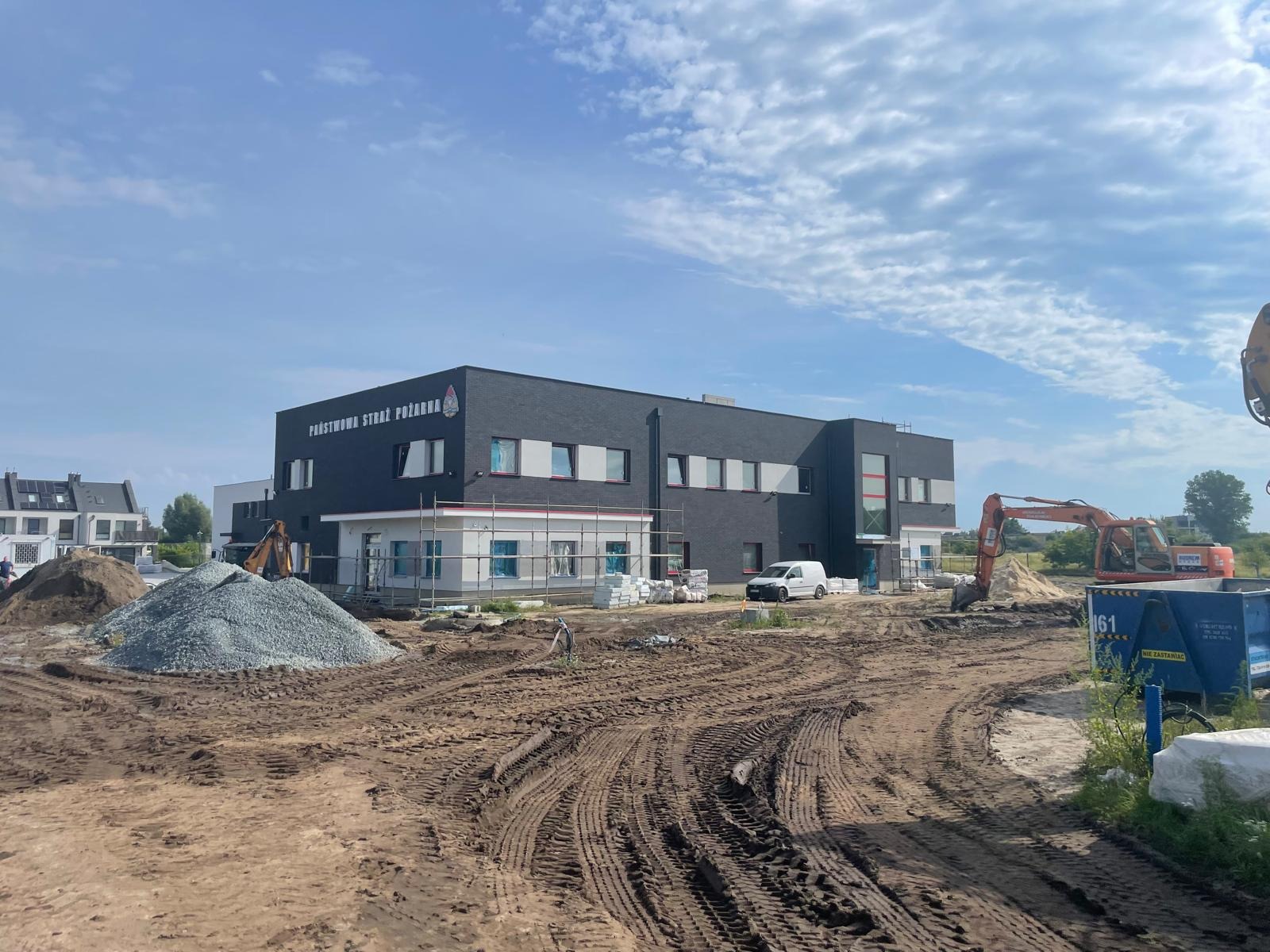 Budynek Państwowej Straży Pożarnej w trakcie budowy, widoczny plac budowy z maszynami, hałda gruzu i częściowo otynkowana elewacja. Słoneczny dzień, błękitne niebo.