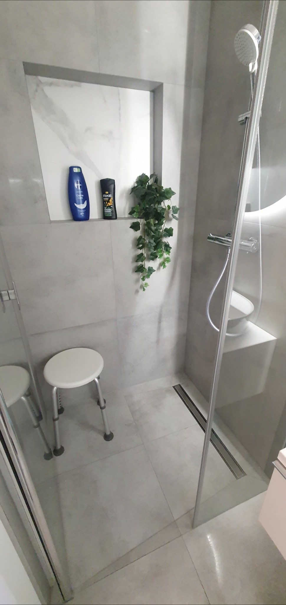 Jasna łazienka z kabiną prysznicową typu walk-in, z siedziskiem, wnęką z kosmetykami i sztuczną rośliną.