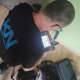 KN Instalacje Kamil Najdek - Elektryk z firmy z Nowogrodu Bobrzańskiego, w koszulce z logo, używa specjalistycznego miernika do pomiaru parametrów instalacji elektrycznej w pomieszczeniu z jasnymi ścianami i drewnopodobną...