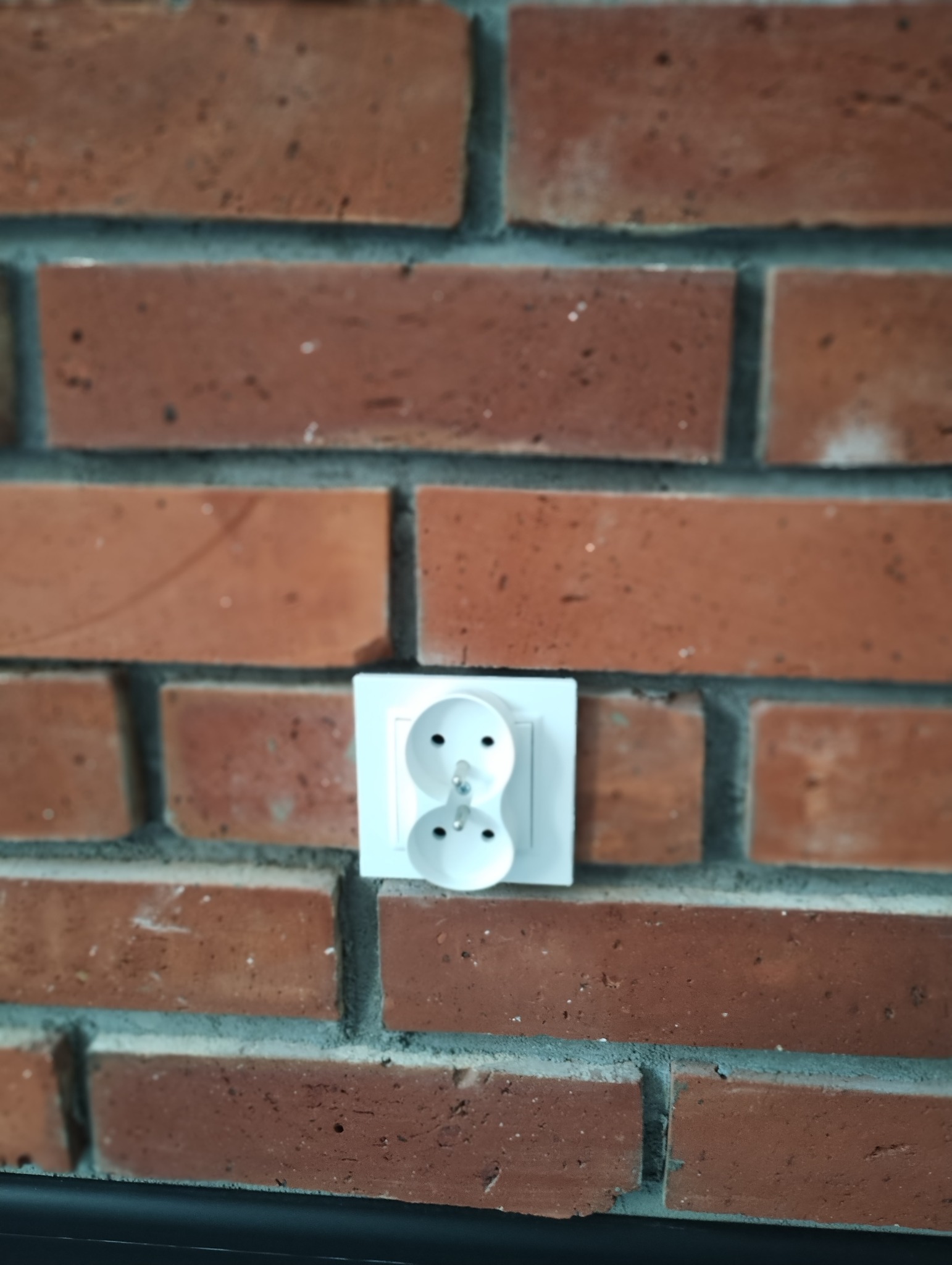 Białe, podwójne gniazdko elektryczne z bolcem, zamontowane na ścianie z czerwonej cegły z szarą fugą.