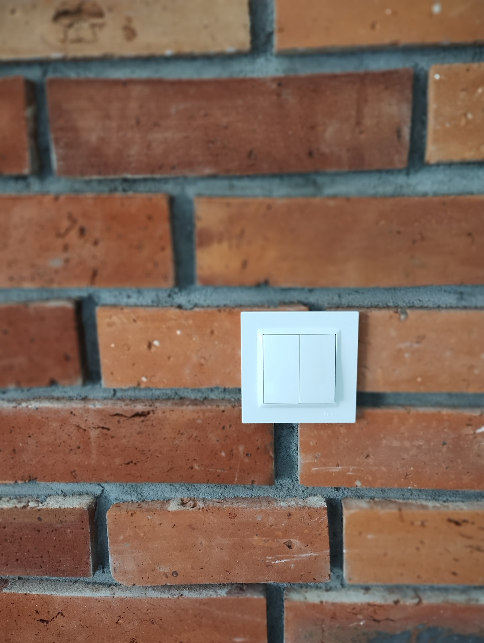 Biały, podwójny włącznik światła marki Simon zamontowany na ścianie z czerwonej cegły dekoracyjnej z szarą fugą.