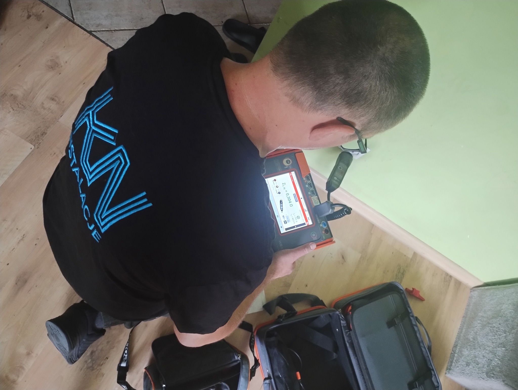 Elektryk z firmy z Nowogrodu Bobrzańskiego, w koszulce z logo, używa specjalistycznego miernika do pomiaru parametrów instalacji elektrycznej w pomieszczeniu z jasnymi ścianami i drewnopodobną...
