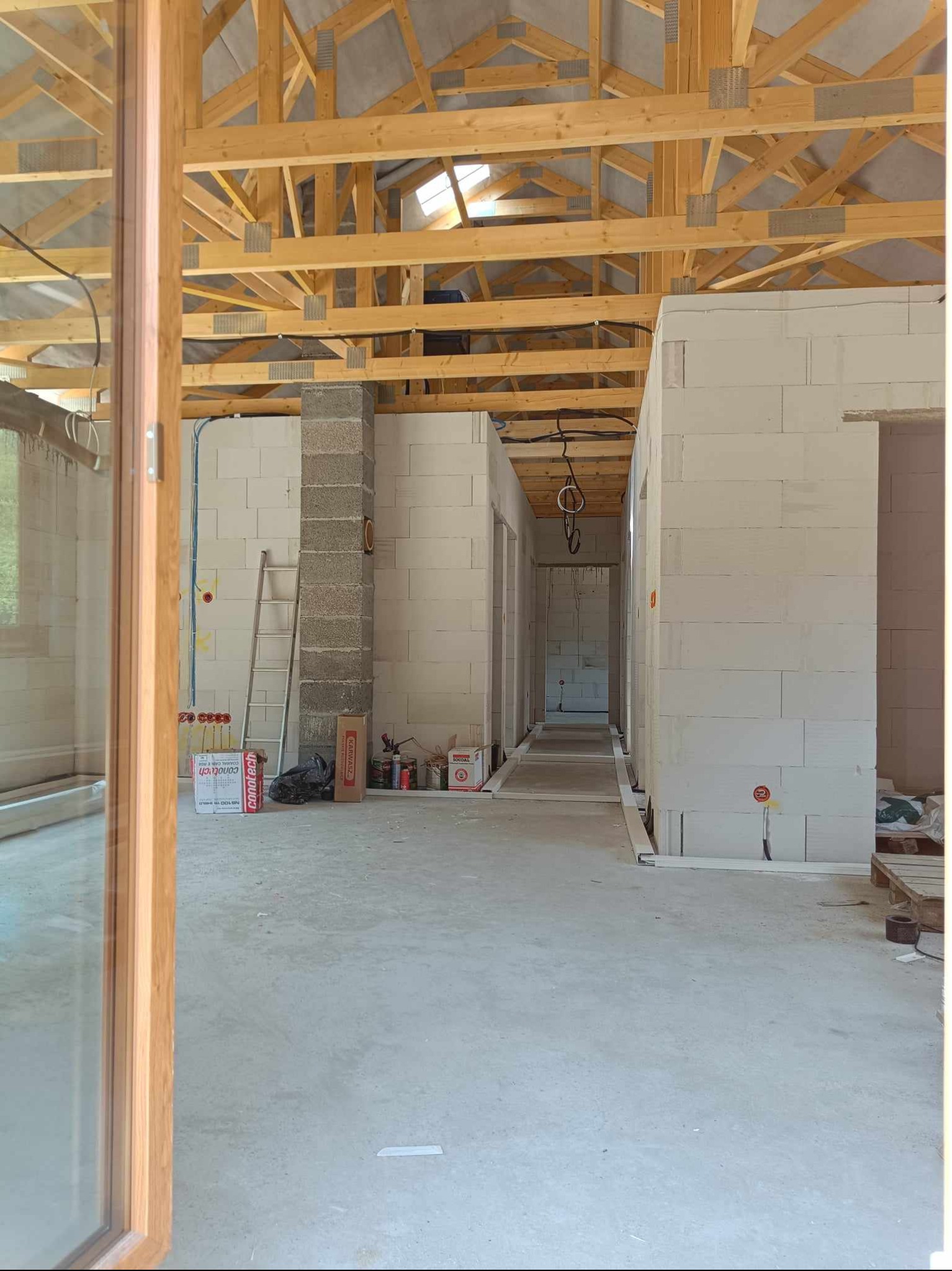 Wnętrze surowego domu w trakcie budowy: widoczne ściany z betonu komórkowego, drewniana konstrukcja dachu, drabina oparta o filar i częściowo poprowadzone przewody elektryczne w ścianach.