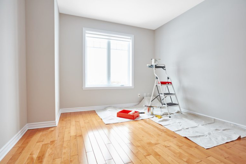 Przygotowania do malowania pokoju: drabina z puszkami farby, kuweta z wałkiem, folia ochronna na parkiecie, świeżo pomalowane ściany na szaro.